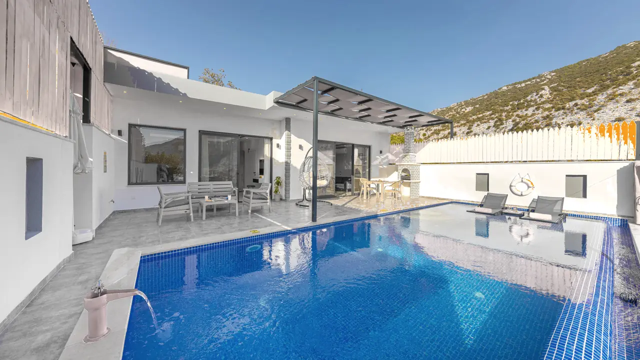 Piscina de gresite azul junto a una casa blanca con porche y tumbonas, a pleno sol