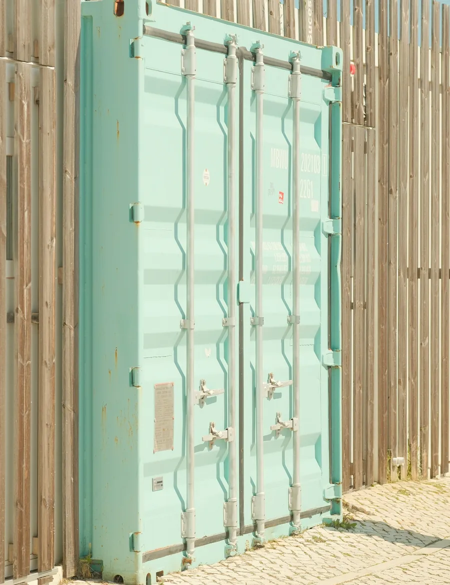 Contenedor marítimo de color verde agua con sus puertas cerradas, junto a una valla de listones de madera