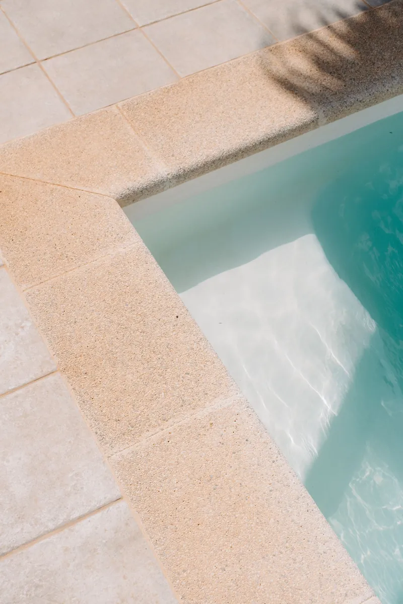 Esquina de una piscina con coronación de piedra artificial de color arena y playa de baldosa clara, con el agua turquesa