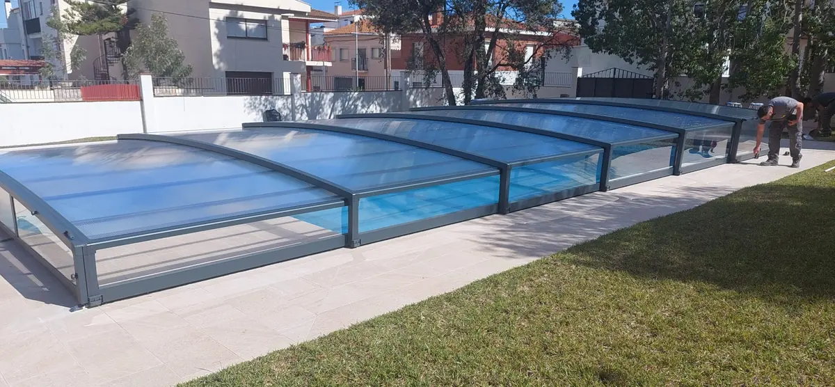 Cubierta telescópica baja de módulos de aluminio antracita y policarbonato celular extendida sobre una piscina rectangular, junto al césped