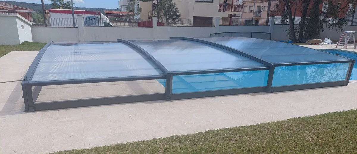 Lateral de una cubierta telescópica baja de aluminio y policarbonato, a poca altura sobre la playa de piedra clara de la piscina