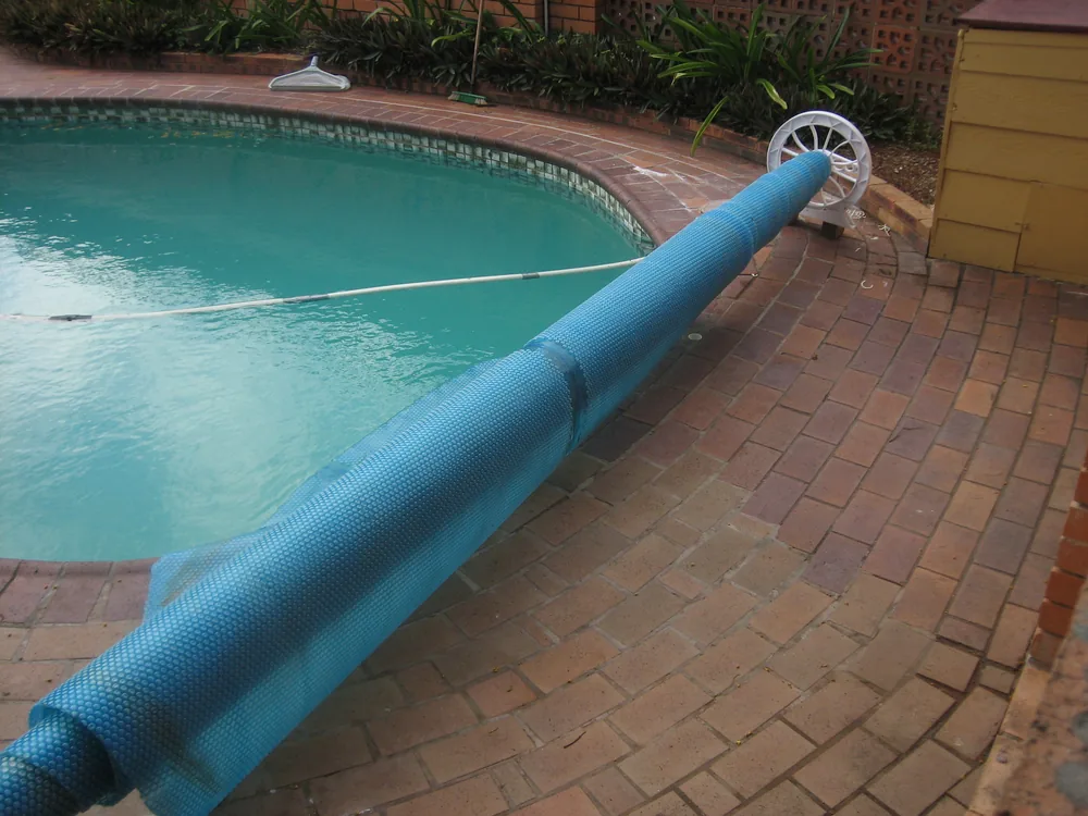 Manta térmica de burbujas enrollada en un enrollador manual con rueda junto a una piscina de obra