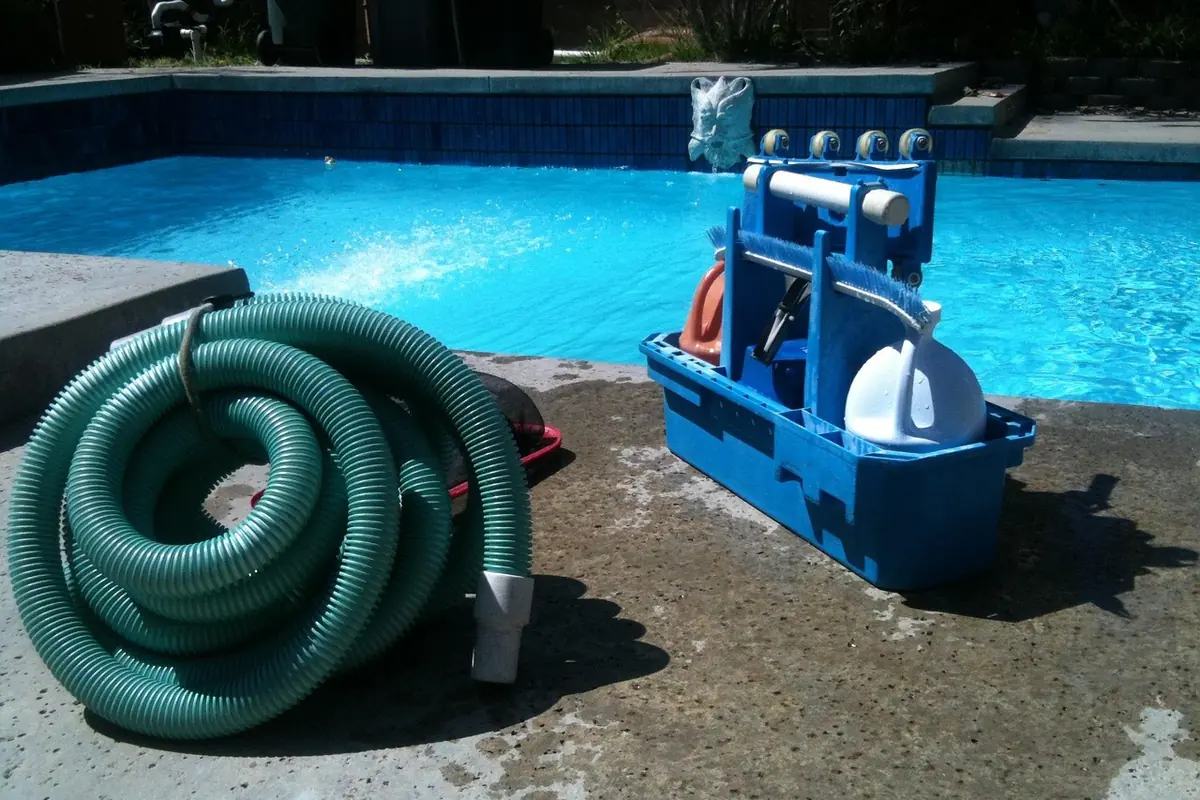 Equipo de aspirado manual de piscina con manguera enrollada y carro con cepillo junto a una piscina