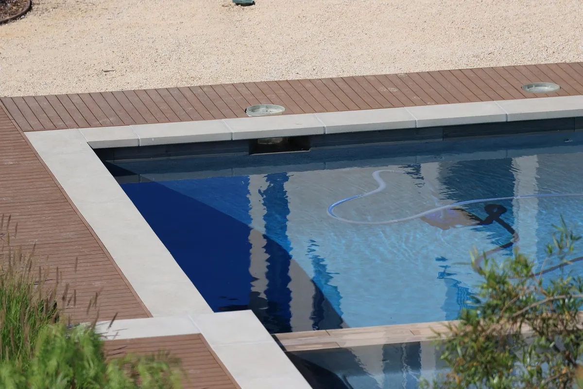 Piscina de jardín con agua azul, coronación clara y un limpiafondos con su manguera trabajando en el fondo