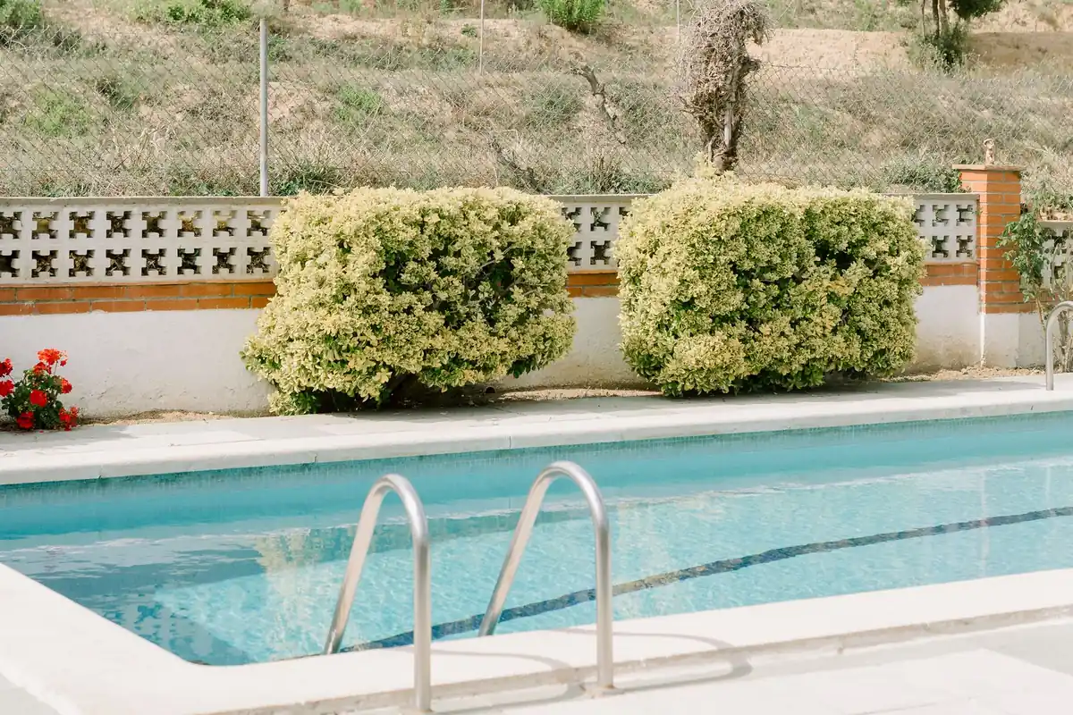 Piscina de obra terminada en un jardín, con coronación de piedra clara, playa perimetral y escalera de acero inoxidable