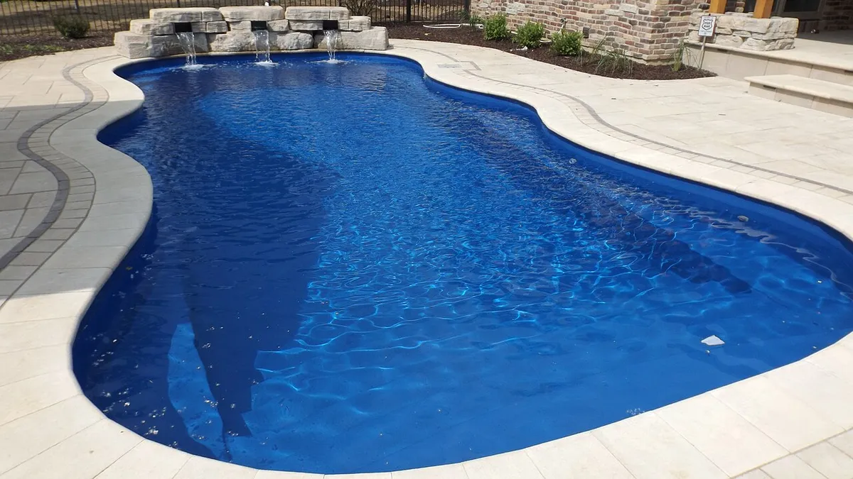 Piscina prefabricada de fibra de vidrio de forma libre, con el vaso azul de una pieza y coronación de piedra clara en un jardín
