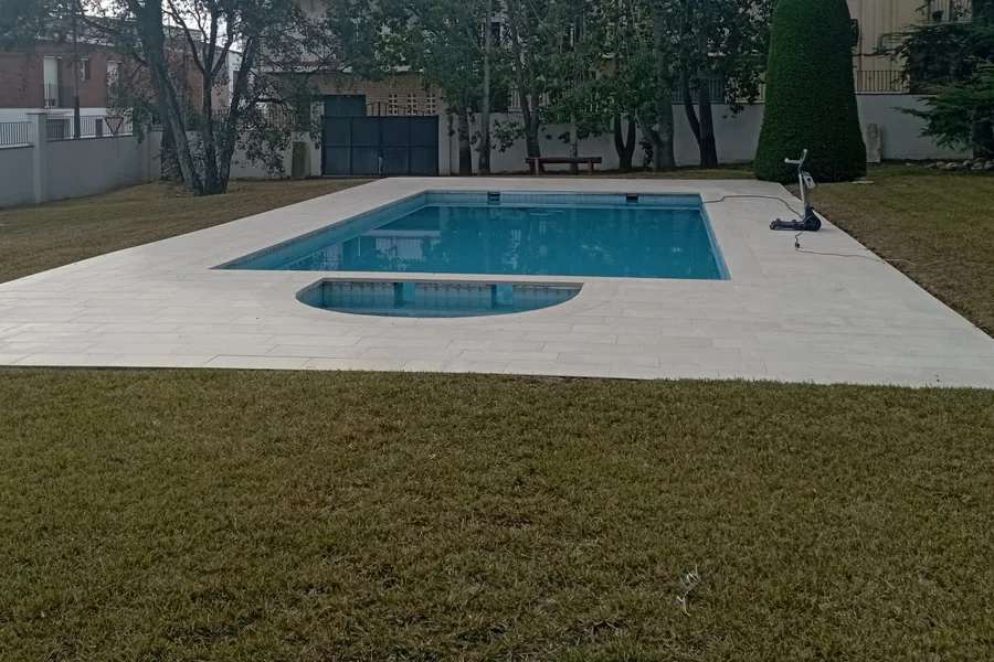 Piscina rectangular con agua limpia, zona semicircular y playa de piedra clara, sin cubierta, en un jardín de césped