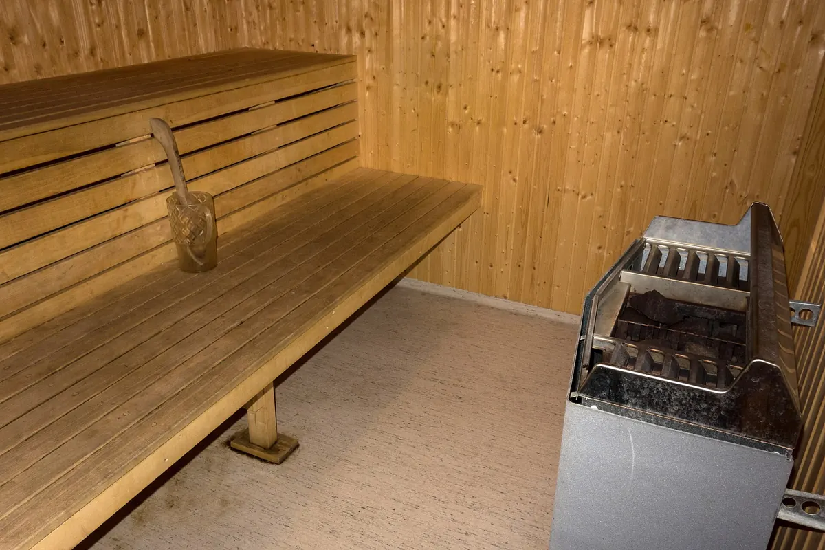 Interior de una sauna con banco de madera de dos alturas, cubo con cazo y estufa eléctrica con piedras